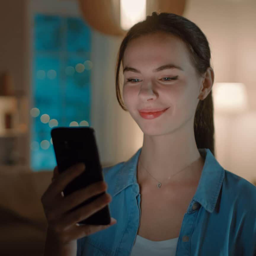 Portrait of Young Beautiful Woman Using Smartphone, Browsing in Internet, Checking Social Networks and Using Smart Home Application while Standing At Home. Cozy Evening.